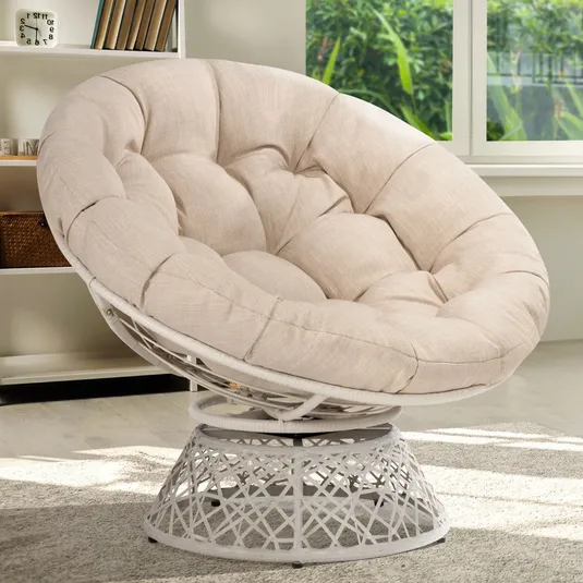 Beige tufted papasan chair with a metal base, placed in a bright room with bookshelves and windows in the background.