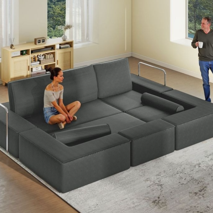 A modular, black fabric sofa featuring adjustable sections and built-in handrails. It creates a large seating area and includes cylindrical armrests. Two people are present, one seated on the sofa and the other standing nearby.