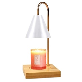 A modern candle warmer lamp with a wooden base, gold metal arm, and cone-shaped white shade, designed to gently heat a decorative pink candle with a detailed label.