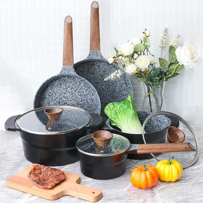 Granite-coated cookware set with 12 pieces, includes two frying pans, a large pot with glass lid, a medium saucepan with lid, and a ladle. The set features wooden handles and a speckled gray finish, accented by a small cutting board with steak and decorative pumpkins.