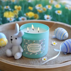 A light blue candle labeled \“Cottontail Dreams\“ with three flames sits on a tray. Nearby are a cute white bunny plush and decorative pastel eggs amidst a floral background.