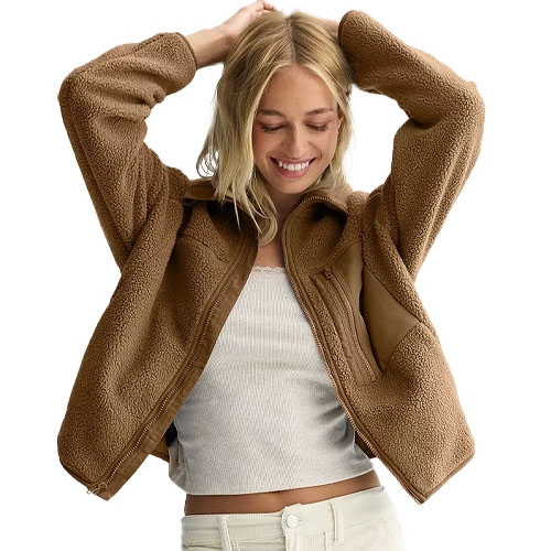 A woman is wearing a brown sherpa-lined fleece jacket over a white ribbed top and light-colored pants, smiling with her hands on her head.