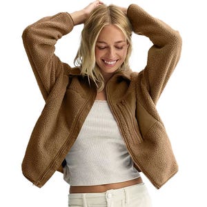 A woman is wearing a brown sherpa-lined fleece jacket over a white ribbed top and light-colored pants, smiling with her hands on her head.