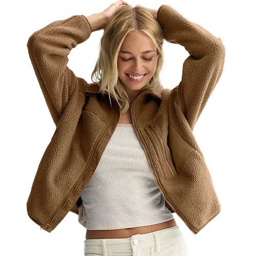 A woman is wearing a brown sherpa-lined fleece jacket over a white ribbed top and light-colored pants, smiling with her hands on her head.