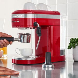 A red KitchenAid espresso machine with a steam wand is shown on a countertop. A hand is positioning a portafilter, and an espresso cup is placed to receive coffee.