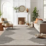 A living room features a beige and black geometric area rug, a cozy cream sofa with orange and gray pillows, a white fireplace with decorative vases, a large leafy plant, and a comfortable beige armchair next to a wicker side table.