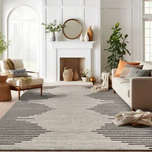 A living room features a beige and black geometric area rug, a cozy cream sofa with orange and gray pillows, a white fireplace with decorative vases, a large leafy plant, and a comfortable beige armchair next to a wicker side table.