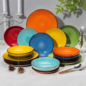 A set of colorful ceramic dinnerware, including plates and bowls in shades of orange, red, yellow, green, blue, and turquoise, arranged on a white surface with decorative elements like candles, pinecones, and a glass bottle.