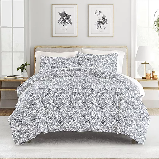 A bed features a grey and white floral patterned comforter with matching pillow shams. The room includes wooden side tables, framed botanical prints, and decorative plants.