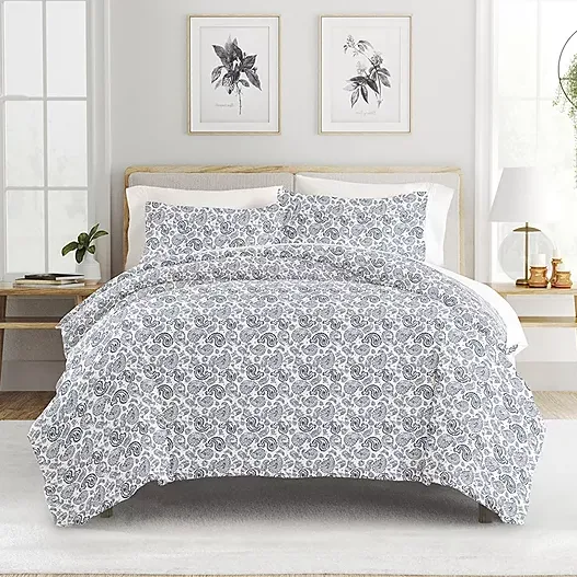 A bed features a grey and white floral patterned comforter with matching pillow shams. The room includes wooden side tables, framed botanical prints, and decorative plants.