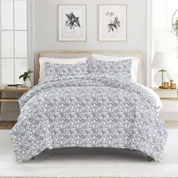 A bed features a grey and white floral patterned comforter with matching pillow shams. The room includes wooden side tables, framed botanical prints, and decorative plants.