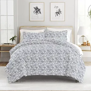 A bed features a grey and white floral patterned comforter with matching pillow shams. The room includes wooden side tables, framed botanical prints, and decorative plants.