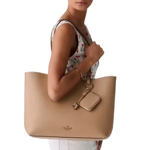 A woman carries a beige designer tote bag with a small attached pouch, wearing a sleeveless top with a cherry pattern.