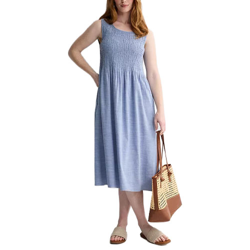 A woman is wearing a light blue, sleeveless, smocked midi dress, holding a brown and beige woven tote bag, and wearing beige slide sandals.