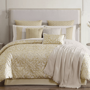 A luxurious bedding set featuring a gold and cream comforter with intricate patterns, matching pillow shams, several decorative throw pillows, and a fringed blanket draped over the side.