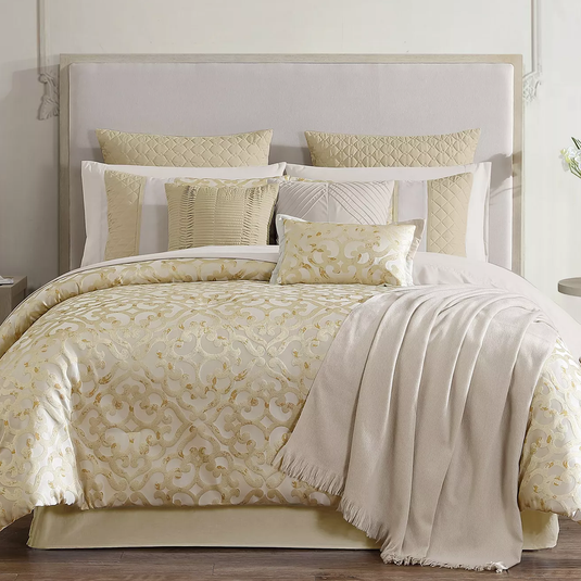 A luxurious bedding set featuring a gold and cream comforter with intricate patterns, matching pillow shams, several decorative throw pillows, and a fringed blanket draped over the side.