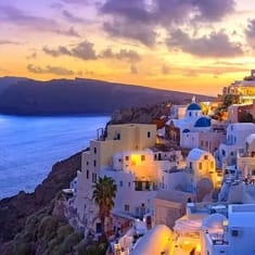 A picturesque view of a coastal town at sunset, featuring whitewashed buildings, blue domed roofs, and a scenic cliffside overlooking the sea.