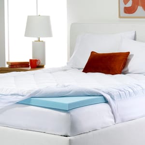 Sealy Memory Foam Topper Sets feature a blue memory foam topper under a plush white quilted cover, providing added comfort and support to the mattress. The setup includes white bedding with a decorative orange pillow, and a side table with a lamp and books.