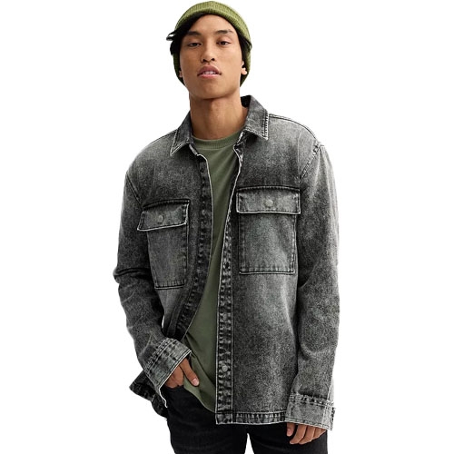 A man is wearing a faded black denim jacket with two chest pockets over an olive green shirt, paired with a green beanie and dark pants.