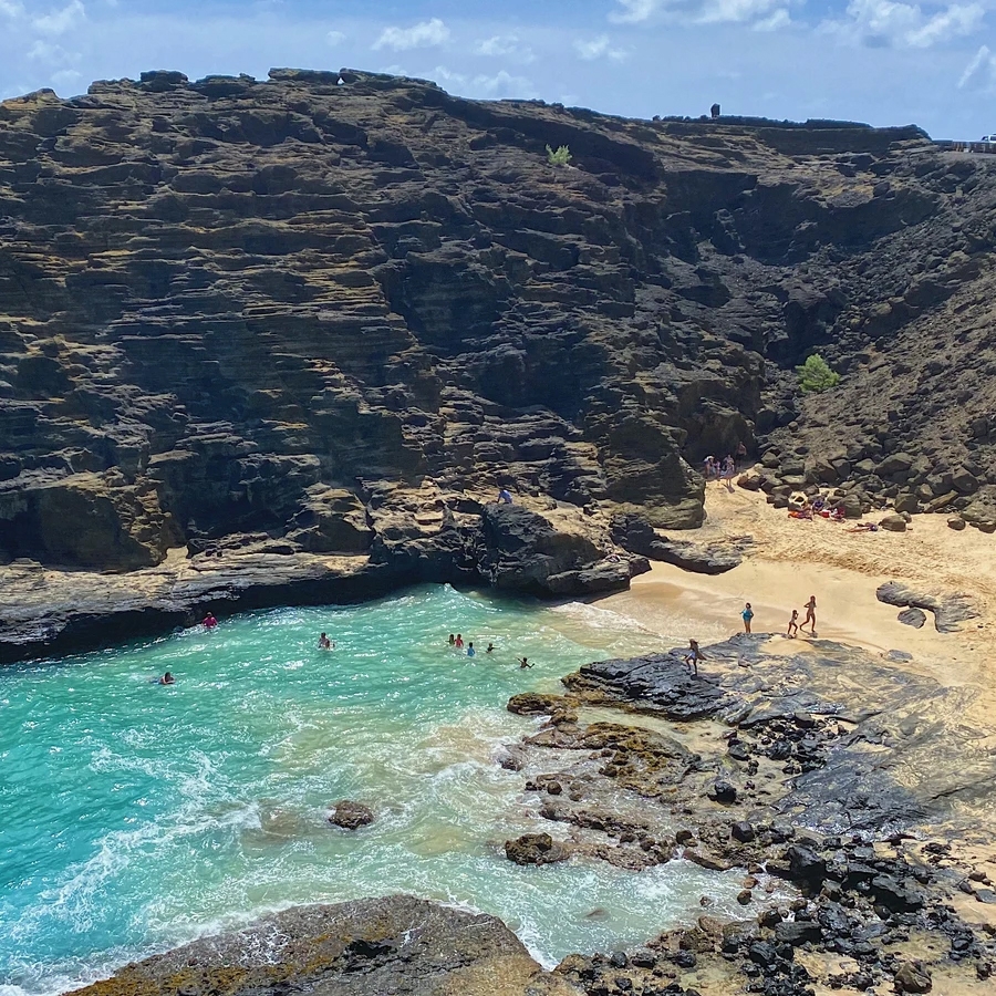 A small beach cove with turquoise waters bordered by rocky cliffs. People are swimming and sunbathing on the sand, surrounded by natural rugged terrain.