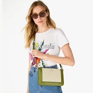 A woman wearing a white T-shirt with colorful print, sunglasses, denim jeans, and carrying a small green crossbody bag.