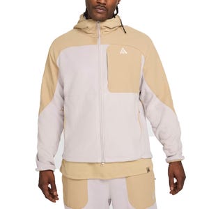 A hooded jacket with a color-block design in beige and light grey, featuring a full zip front and a chest pocket with a small triangular logo. It is paired with matching pants that have similar color blocking.