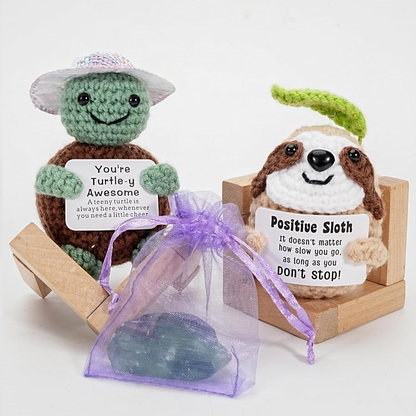 Two crochet figures: a turtle with a hat holding a sign saying \“You're Turtle-y Awesome,\“ and a sloth with a leaf on its head holding a sign saying \“Positive Sloth.\“ A purple mesh bag with a stone is positioned in front.