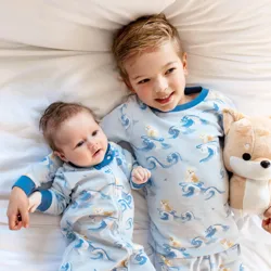 Two children, a baby and a young boy, wear matching blue pajamas featuring surfing animals. The boy holds a plush toy resembling a fox.