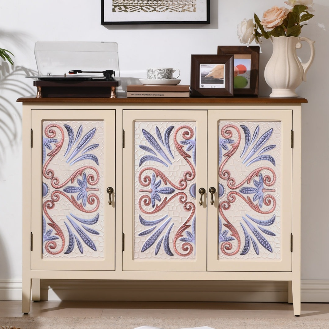 A beige cabinet with floral-patterned doors holds a record player, decorative items, and a vase with flowers on top. A framed wall art piece hangs above, and an open magazine lies on the floor nearby.