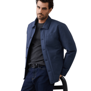 A man is wearing a navy blue jacket over a dark sweater and jeans with a black belt, leaning against a chair with a neutral expression.
