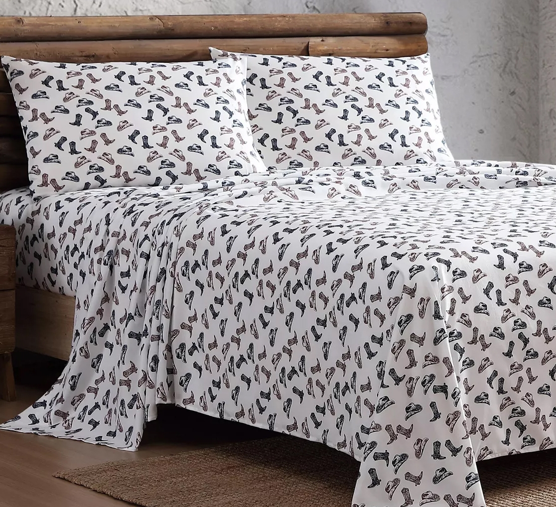 A bed is covered with a sheet and pillowcases featuring a pattern of various boots and shoes. The bedding has a rustic theme with a log frame in the background.