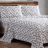 A bed is covered with a sheet and pillowcases featuring a pattern of various boots and shoes. The bedding has a rustic theme with a log frame in the background.