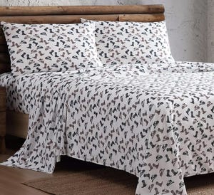 A bed is covered with a sheet and pillowcases featuring a pattern of various boots and shoes. The bedding has a rustic theme with a log frame in the background.