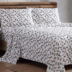 A bed is covered with a sheet and pillowcases featuring a pattern of various boots and shoes. The bedding has a rustic theme with a log frame in the background.