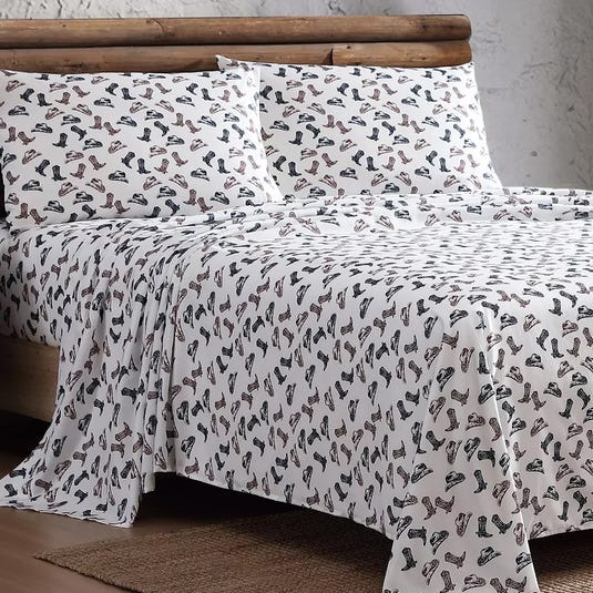 A bed is covered with a sheet and pillowcases featuring a pattern of various boots and shoes. The bedding has a rustic theme with a log frame in the background.