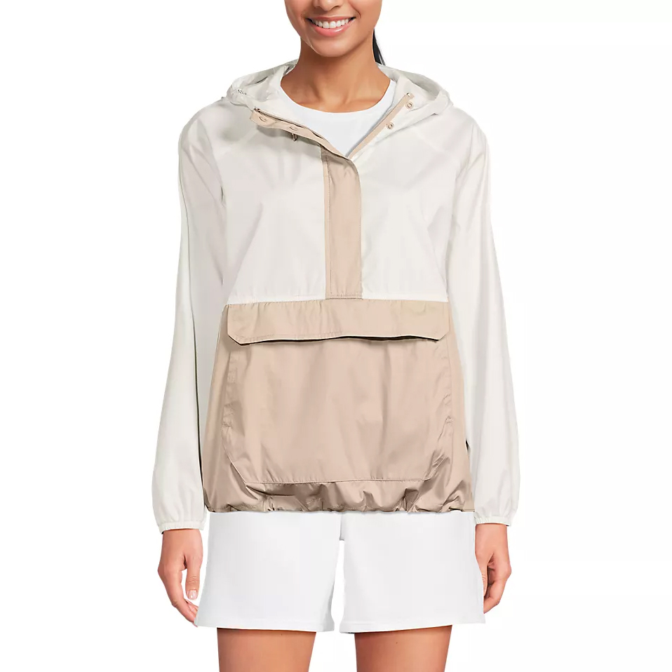 A woman is wearing a color-blocked hooded windbreaker with a large front pocket. The jacket is beige and white, paired with white shorts.
