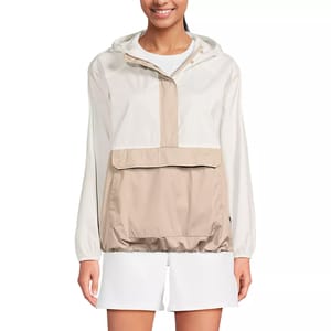 A woman is wearing a color-blocked hooded windbreaker with a large front pocket. The jacket is beige and white, paired with white shorts.