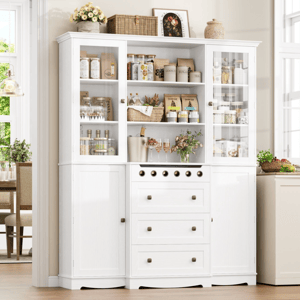 White kitchen cabinet with glass doors displaying jars, baskets, and a wine rack. It features drawers and cupboards below, and decorative items like flowers and framed art above.