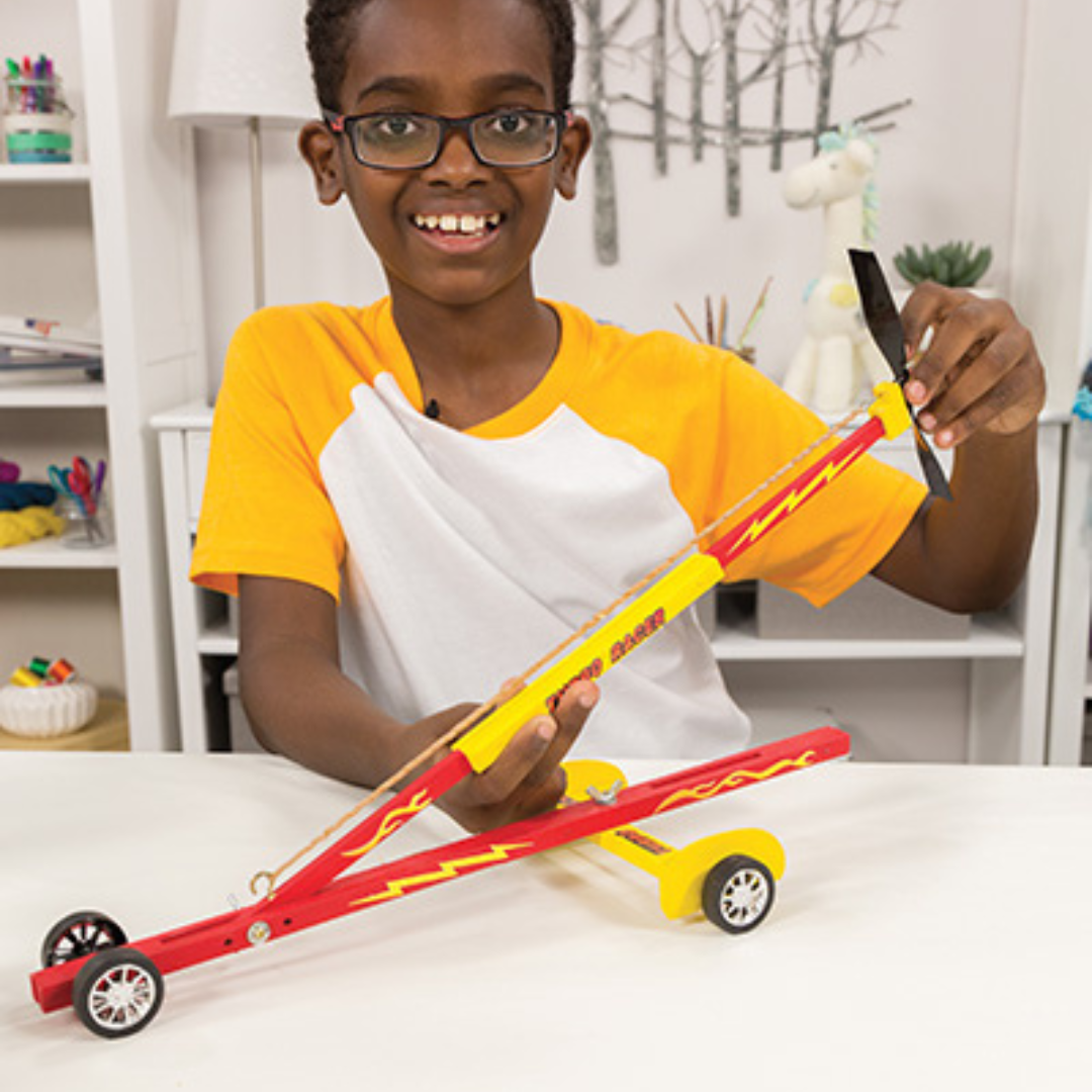 A smiling child is holding a model red and yellow racing car with a long body and large rear wheels.