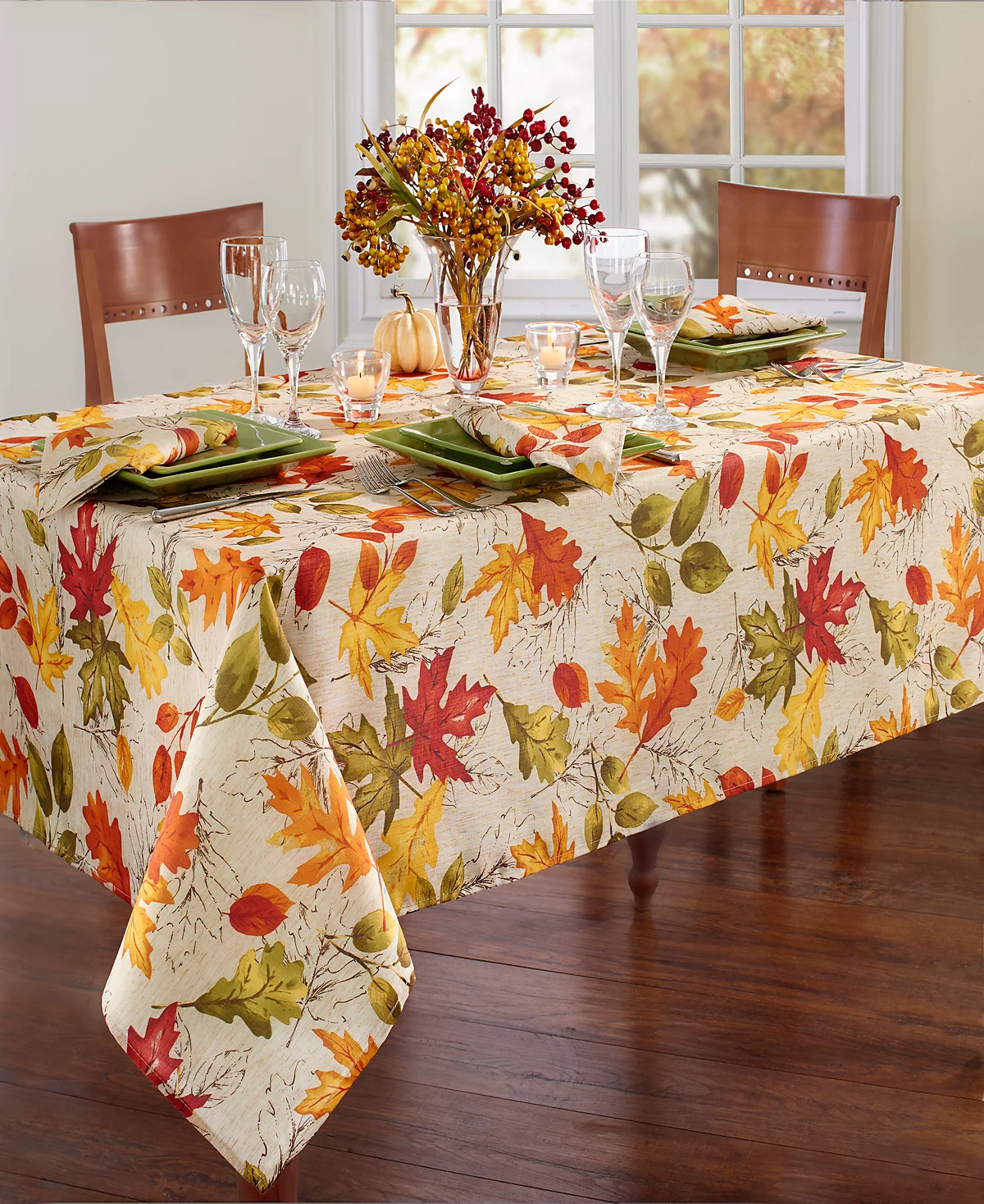 A fall-themed table setting features a tablecloth with colorful autumn leaves, green square plates, wine glasses, and a centerpiece of seasonal foliage and small pumpkins.