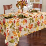 A fall-themed table setting features a tablecloth with colorful autumn leaves, green square plates, wine glasses, and a centerpiece of seasonal foliage and small pumpkins.
