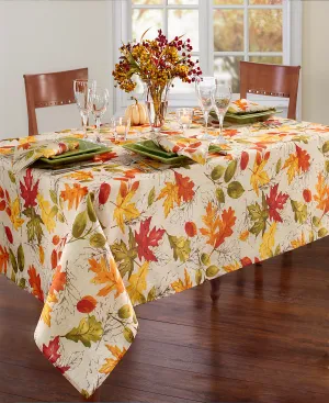 A fall-themed table setting features a tablecloth with colorful autumn leaves, green square plates, wine glasses, and a centerpiece of seasonal foliage and small pumpkins.