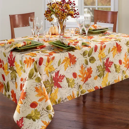 A fall-themed table setting features a tablecloth with colorful autumn leaves, green square plates, wine glasses, and a centerpiece of seasonal foliage and small pumpkins.