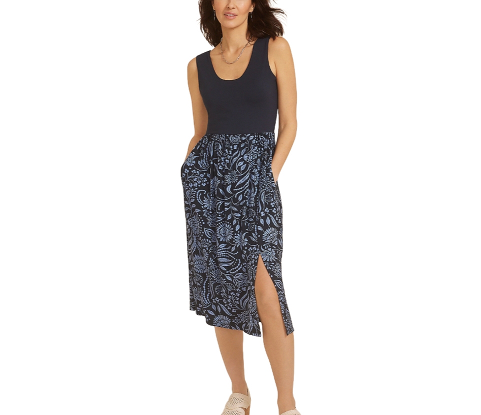 A woman is wearing a sleeveless dress with a solid black top and a blue floral-patterned skirt featuring a side slit, paired with light-colored woven shoes.