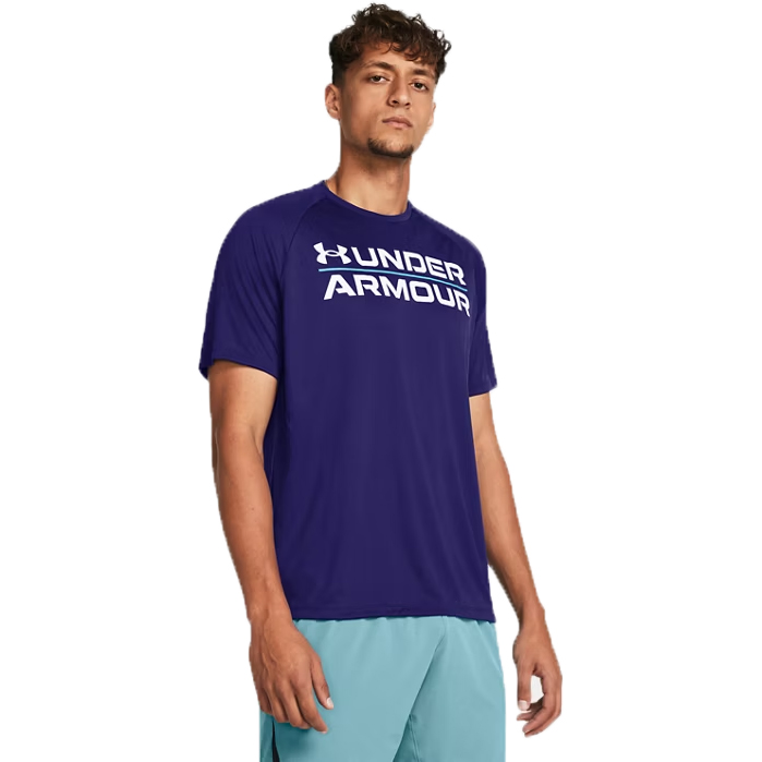 The UA Velocity Graphic Short Sleeve Crew is a men's athletic shirt in deep purple, featuring the \“Under Armour\“ logo in bold white across the chest. It's paired with light blue shorts, offering a sporty and casual look.