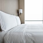 A neatly made bed with white sheets and pillows beside a window in a minimalist bedroom.