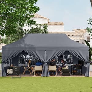 A large gray outdoor canopy tent is set up on a patio with people sitting inside on chairs, surrounded by tables with beverages and items. The scene is shaded by trees, and a house with tiled roofs is in the background.