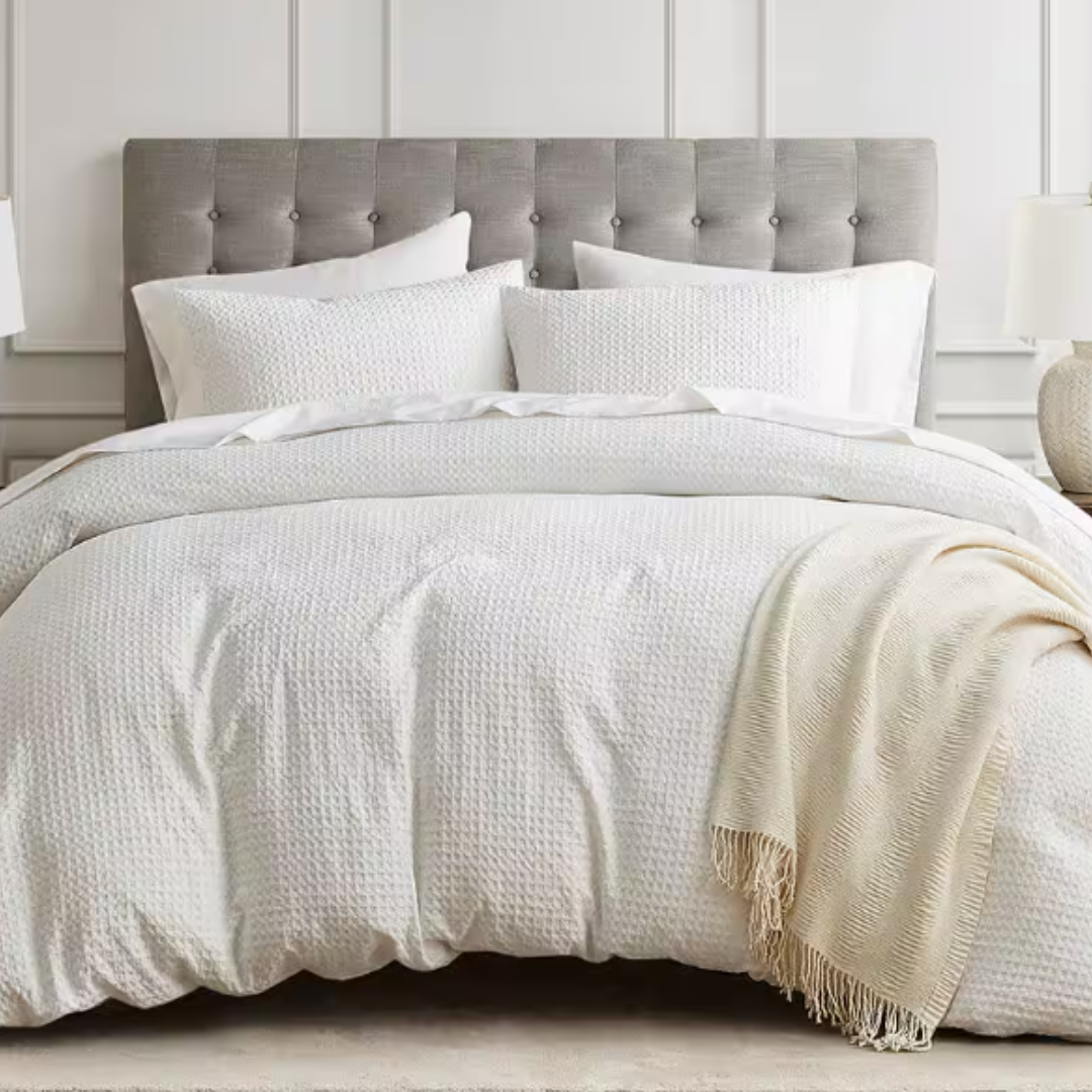 Gray tufted headboard with a 3-piece white waffle weave duvet cover set, including textured duvet and pillow shams, accompanied by a light beige throw. Room decor features neutral tones and a modern, elegant design.