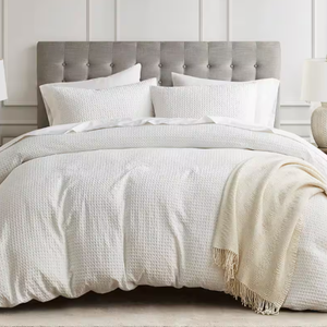 Gray tufted headboard with a 3-piece white waffle weave duvet cover set, including textured duvet and pillow shams, accompanied by a light beige throw. Room decor features neutral tones and a modern, elegant design.