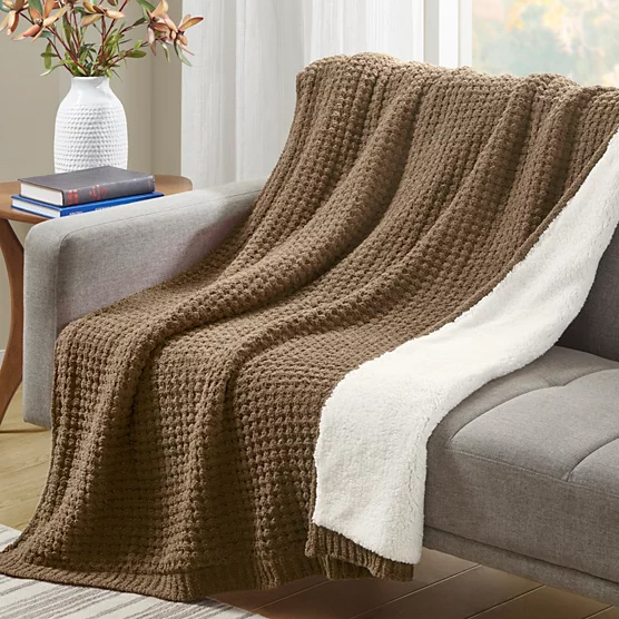 A plush, reversible throw with a brown chenille front and a white sherpa back, draped over a gray couch, accompanied by a vase and books on a wooden side table.